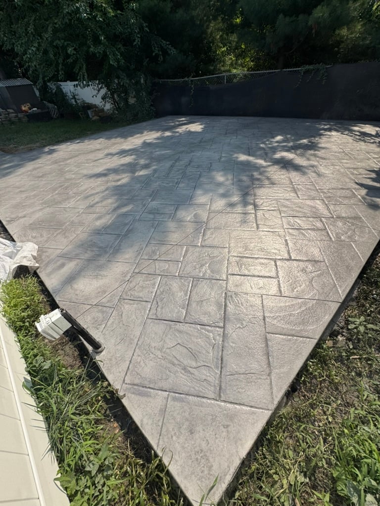 Newly installed gray stamped concrete patio with geometric brick pattern surrounded by green lawn and black fence