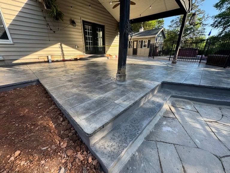 Concrete patio with pergola structure attached to a beige house with landscaping and trees visible