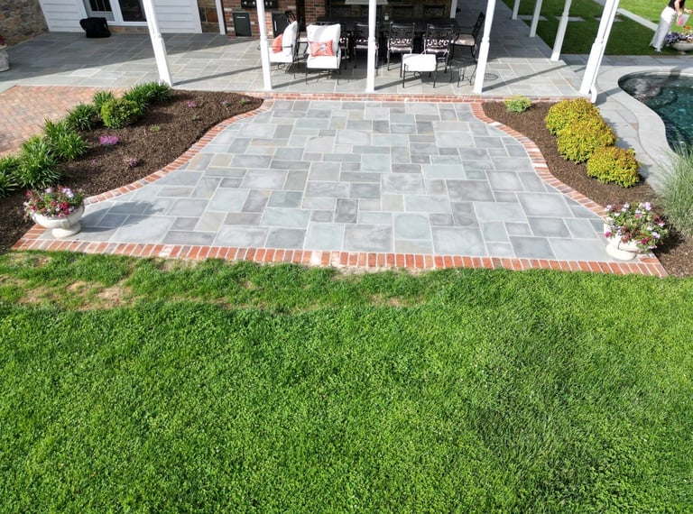 Paved patio with herringbone pattern pavers edged in red brick, flanked by landscaping beds and green lawn in front of a house with white columns
