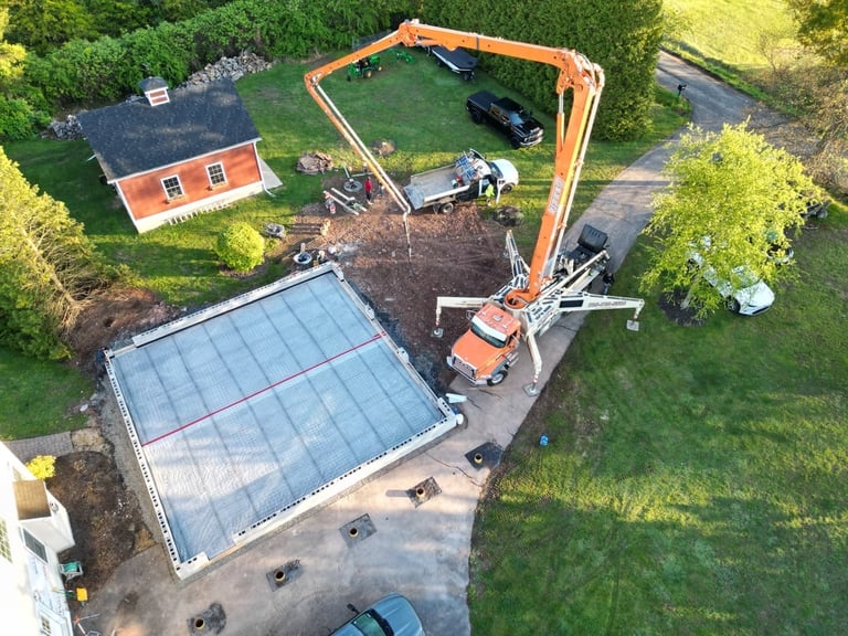 Aerial view of construction site with orange excavator, pool foundation, residential property, and landscaping surrounded by green grass and trees