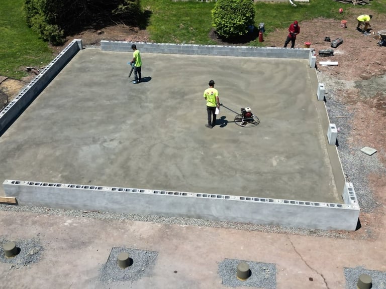 Aerial view of construction workers in yellow safety vests finishing a concrete foundation, surrounded by green landscaping.