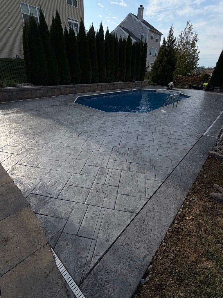 Residential backyard with in-ground swimming pool surrounded by gray paver patio, tall evergreen shrubs, and a house in the background