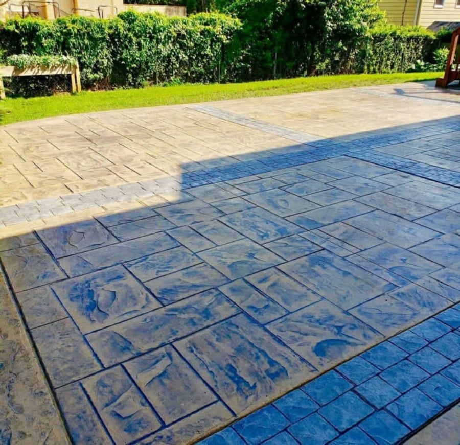Patio with blue and tan patterned pavers casting afternoon shadows, bordered by green lawn and hedges