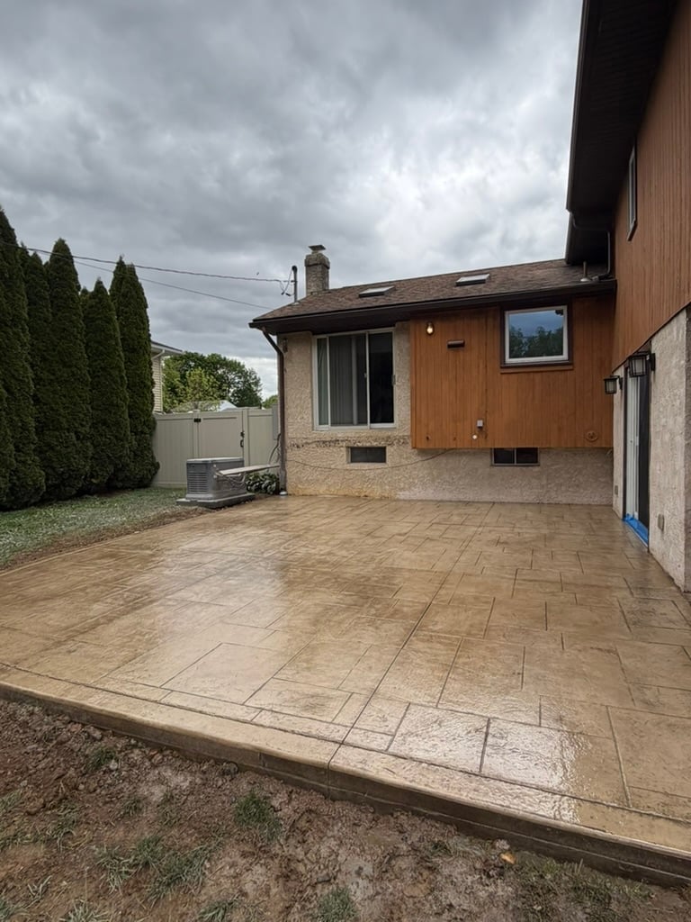 Residential backyard with stamped concrete patio, brick house with wood siding, green hedges, and overcast sky