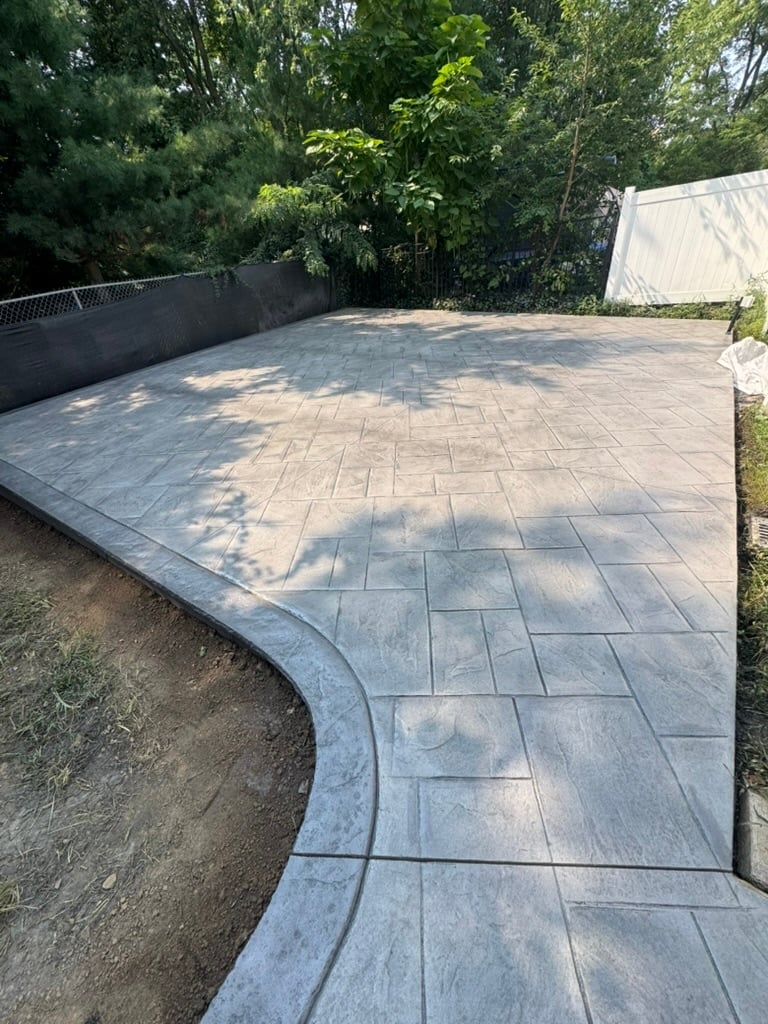 Paved patio with curved herringbone pattern pavers, black fence, and dense green trees in background.