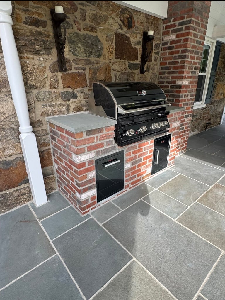 Outdoor kitchen with built-in black gas grill and oven in a brick and stone enclosure with slate flooring