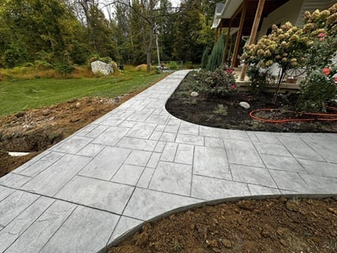 Curved gray concrete paver pathway leading to a house entrance with landscaped garden beds and green lawn
